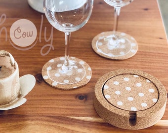 May include: Cork coasters with white polka dots, displayed with wine glasses and a candle on a wooden surface. The coasters are in a holder, and some are under the wine glasses. The image has the text "holy cow".
