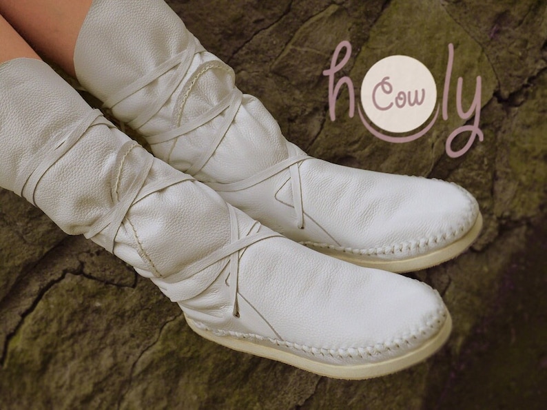 May include: White leather ankle boots with laces tied up the leg. The boots have a stitched seam around the sole. The boots are on a brown textured surface. The text "holy cow" is visible in the background.