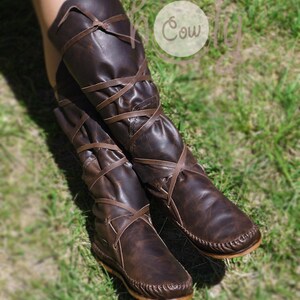 K&ouml;nnte beinhalten: Braune Lederstiefel mit Schn&uuml;rsenkeln, die sich um Kn&ouml;chel und Wade wickeln. Die Stiefel haben eine Mokassin-Spitze und stehen auf einem Bett aus gr&uuml;nem Gras.