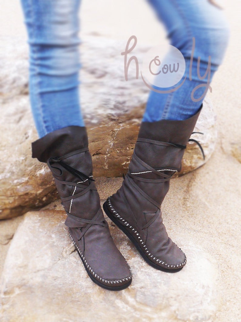 May include: A pair of brown leather boots with a unique design. The boots have a folded-over top, wrap-around laces, and white stitching details. The boots are worn with blue jeans.