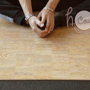 Op de afbeelding: Een rechthoekige yogamat van kurk met een lichtbruin houtnerfpatroon. De mat ligt op een donkergrijze ondergrond. De afbeelding bevat de tekst "holy cow" in een cirkel.