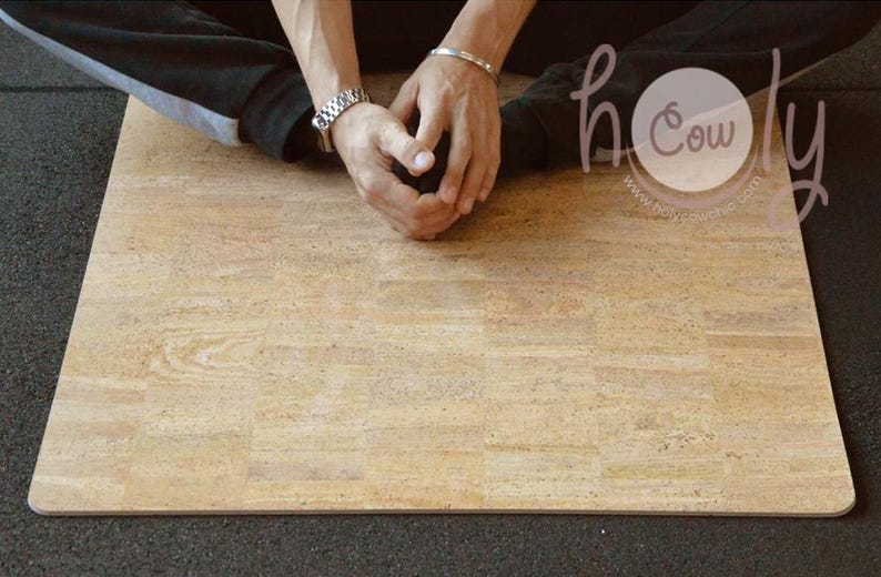 Op de afbeelding: Een rechthoekige yogamat van kurk met een lichtbruin houtnerfpatroon. De mat ligt op een donkergrijze ondergrond. De afbeelding bevat de tekst "holy cow" in een cirkel.