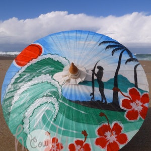 Op de afbeelding: Een kleurrijke strandthema papieren paraplu met een geschilderd tafereel van een surfer, golven, palmbomen en hibiscus bloemen. De paraplu is blauw, groen, rood en wit.