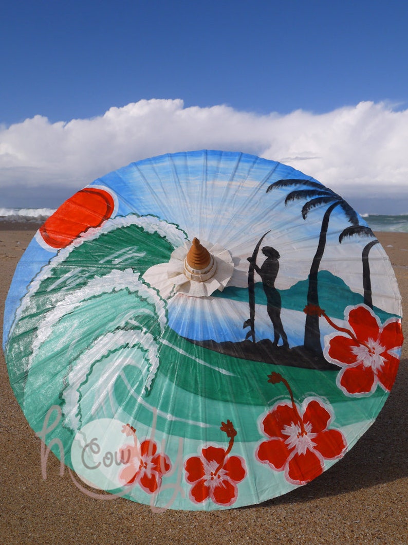 Op de afbeelding: Een kleurrijke strandthema papieren paraplu met een geschilderd tafereel van een surfer, golven, palmbomen en hibiscus bloemen. De paraplu is blauw, groen, rood en wit.