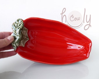 May include: A glossy red ceramic serving dish shaped like a pepper. The dish has a green and brown leaf and stem detail at one end. The dish is held by a hand.