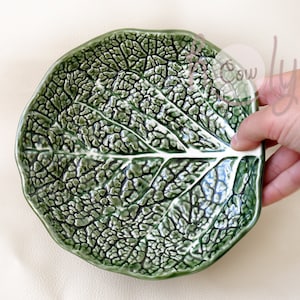 May include: A green ceramic plate shaped like a leaf, featuring a detailed, textured surface that mimics the veins of a cabbage leaf. The plate is held by a hand, showcasing its unique, naturalistic design.
