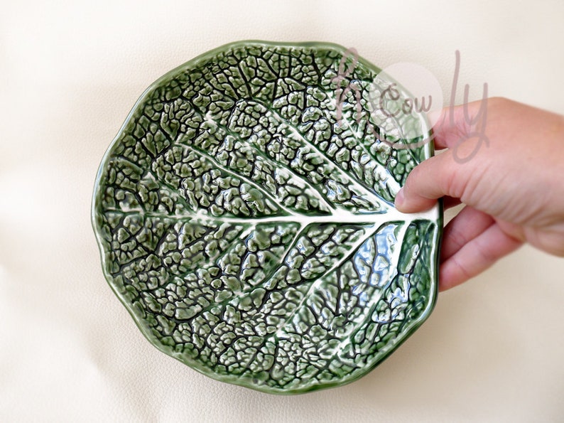 May include: A green ceramic plate shaped like a leaf, featuring a detailed, textured surface that mimics the veins of a cabbage leaf. The plate is held by a hand, showcasing its unique, naturalistic design.
