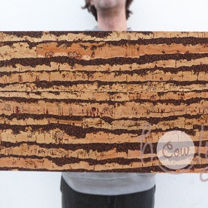 Può includere: Una tavola di sughero rettangolare con un aspetto naturale e strutturato. La tavola presenta bande orizzontali marrone scuro e beige, creando un motivo a righe. Il materiale in sughero è visibile, mostrando la sua unica venatura e variazioni di colore. La tavola è tenuta da una persona.
