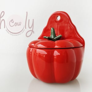 Puede incluir: Recipiente de pared de cerámica roja en forma de tomate con tapa. El recipiente tiene un tallo y una hoja verdes.