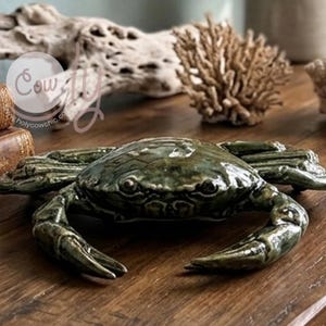 Puede incluir: Una figura de cangrejo de cerámica en color verde oscuro brillante con textura detallada en el caparazón y pinzas extendidas. El cangrejo presenta una forma realista con un acabado liso y reflectante, mostrando detalles moldeados.