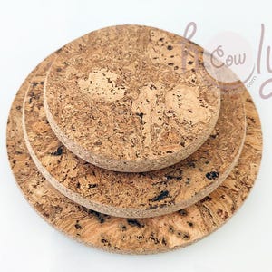 Puede incluir: Tres posavasos redondos de corcho apilados uno encima del otro. Los posavasos están hechos de corcho natural y tienen un color marrón con un patrón moteado.