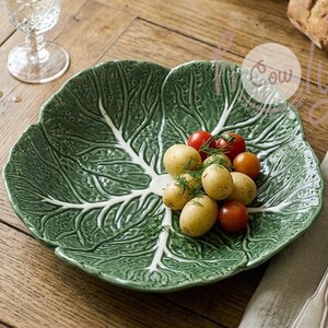 Może przedstawiać: Zielony ceramiczny talerz do serwowania w kształcie liścia kapusty z białymi żyłkami. Zawiera czerwone pomidory i żółte ziemniaki. Na drewnianym stole leżą widelec i nóż.