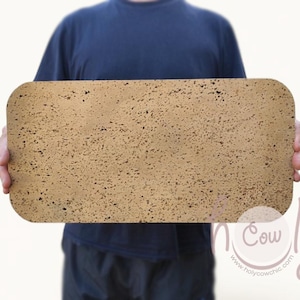 May include: A rectangular cork board with rounded corners is held by a person. The board has a natural, speckled appearance with a light brown color. The background is plain white, and the person is wearing a dark blue shirt.