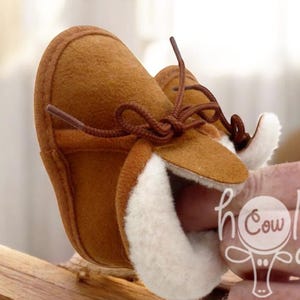 May include: A pair of brown baby moccasins with white faux fur lining and brown laces. The shoes are made of a soft material and are designed for infants. The moccasins are a warm, cozy style.