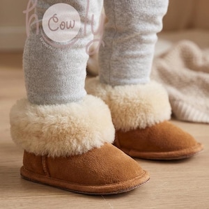 May include: Brown suede baby booties with a cream-colored shearling cuff. The booties are worn with gray leggings. The image is taken from a low angle, showing the booties on a wooden floor.