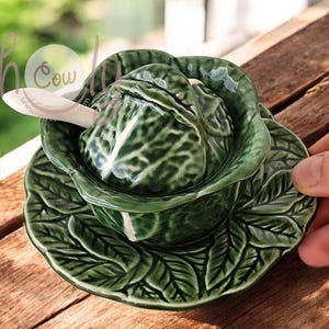 May include: A green ceramic cabbage-shaped serving dish with a matching lid and saucer. The dish includes a small white spoon. The design features detailed leaf patterns. The set is placed on a wooden surface.