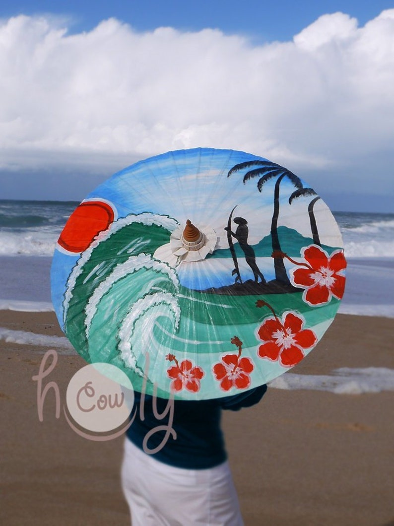Op de afbeelding: Een kleurrijke strandparasol met een golf, palmbomen en hibiscusbloemen erop geschilderd. De parasol is open en wordt door iemand op een strand vastgehouden.