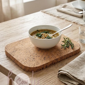 Pode incluir: Um lugar americano retangular de cortiça com cantos arredondados e uma textura natural castanha salpicada. Uma tigela de cerâmica branca com sopa de lentilhas e uma colher de prata está colocada em cima, com um ramo de ervas frescas.