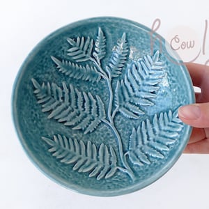Pode incluir: Uma tigela de cerâmica azul-esverdeada com um design de samambaia. A tigela é estampada com um padrão de samambaia e tem uma superfície texturizada.