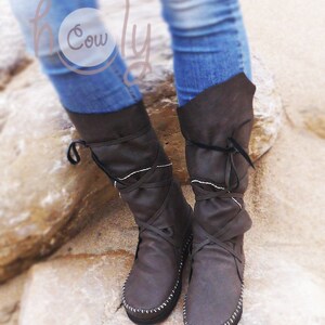 May include: Brown leather knee-high boots with crisscross laces and white stitching. The boots have a folded-over top and are worn with blue jeans. The boots are on a stone surface.