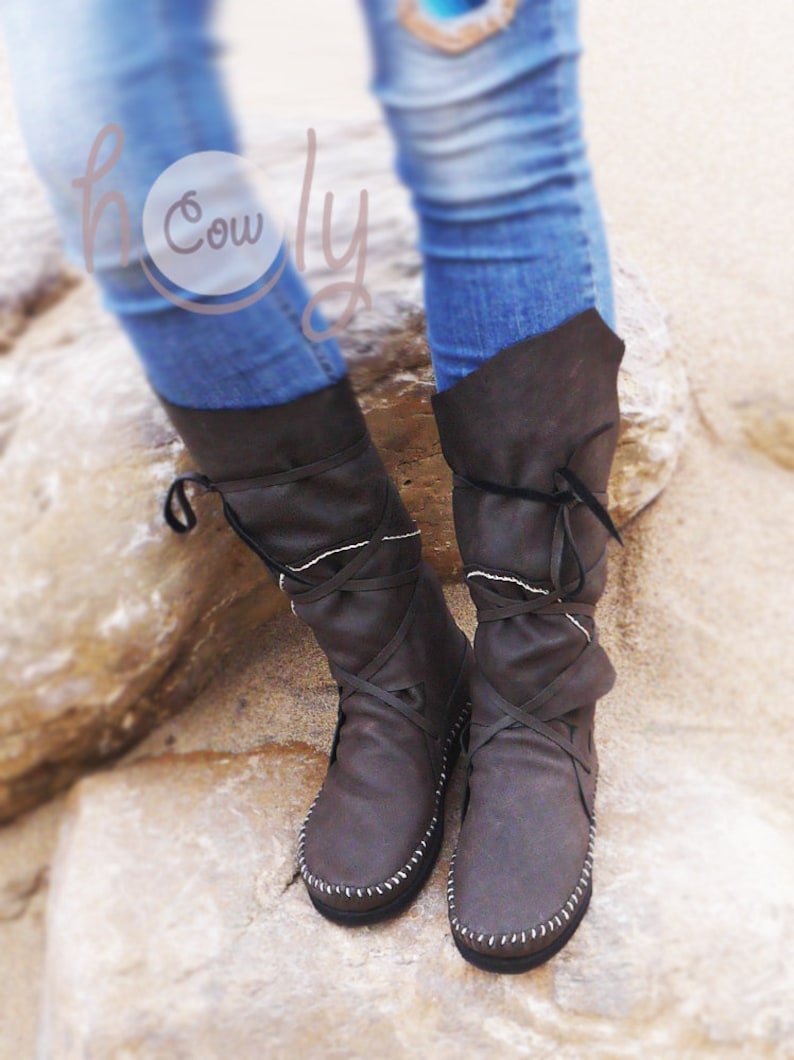 May include: Brown leather knee-high boots with crisscross laces and white stitching. The boots have a folded-over top and are worn with blue jeans. The boots are on a stone surface.