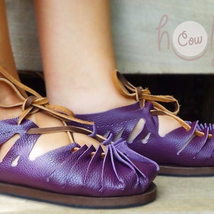 May include: Purple leather sandals with brown laces and cutouts. The sandals have a rounded toe and a brown sole. The sandals are tied at the ankle.