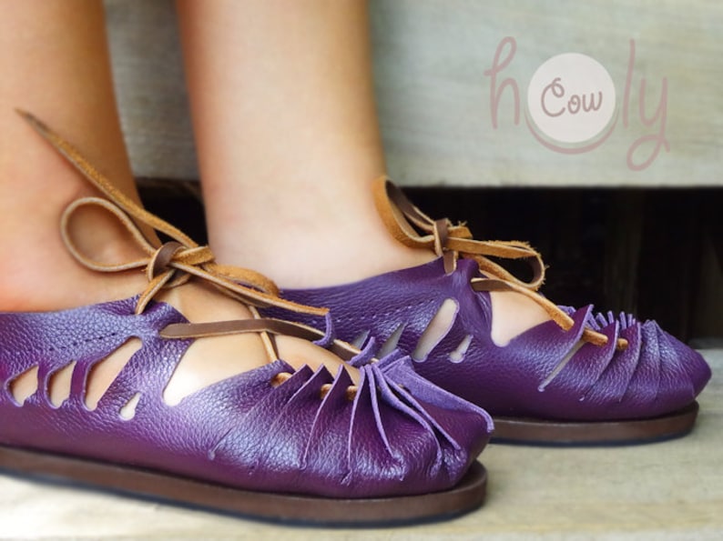 May include: Purple leather sandals with brown laces and cutouts. The sandals have a rounded toe and a brown sole. The sandals are tied at the ankle.