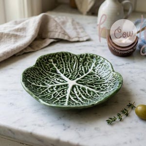 Puede incluir: Un plato de cerámica verde con forma de hoja de col, con vetas detalladas. El plato está sobre una superficie de mármol, con un paño de lino y un pequeño cuenco de madera en el fondo. Una aceituna y una ramita de tomillo están cerca.