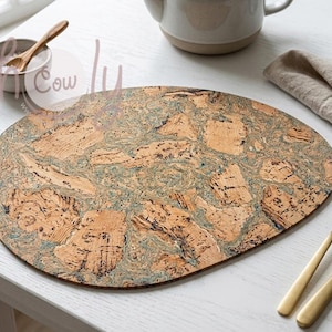 May include: Oval cork placemat with a natural wood grain pattern and blue-green accents. The placemat is on a white table, with gold-colored silverware, a small bowl, and a teapot in the background. The placemat measures approximately 16 inches long.