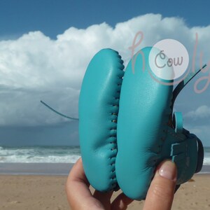 May include: A pair of turquoise blue leather baby shoes with laces. The shoes are held in a hand against a background of a beach and ocean.