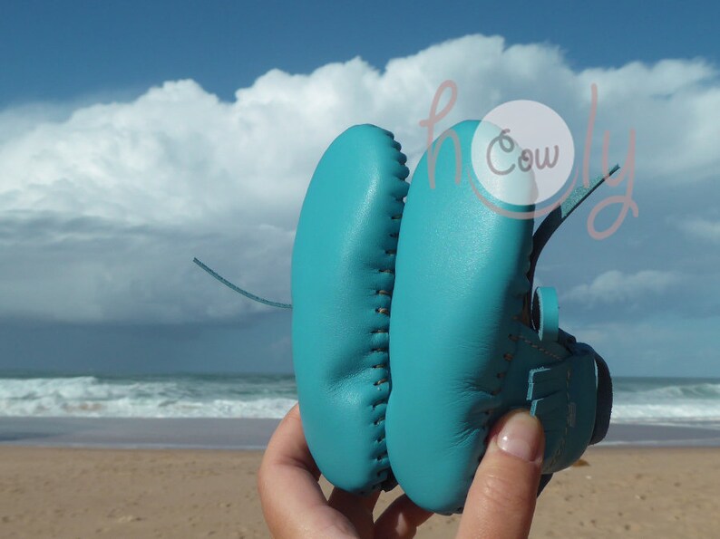 May include: A pair of turquoise blue leather baby shoes with laces. The shoes are held in a hand against a background of a beach and ocean.