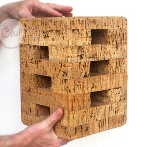 May include: A square-shaped, brown cork block with multiple rectangular holes cut into it. The block is made of natural cork and has a rough, textured surface.
