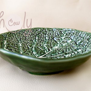 May include: Green ceramic leaf-shaped bowl. The bowl has a textured surface that mimics the veins of a leaf. The bowl is a deep green colour with a glossy finish. The bowl is a decorative piece.