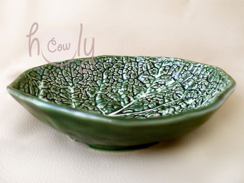 May include: Green ceramic leaf-shaped bowl. The bowl has a textured surface that mimics the veins of a leaf. The bowl is a deep green colour with a glossy finish. The bowl is a decorative piece.
