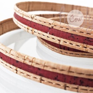 Puede incluir: Primer plano de dos correas de corcho con una franja roja central. Las correas están hechas de material de corcho natural, con costuras visibles a lo largo de los bordes. La franja roja añade un toque de color al corcho neutro.