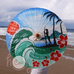 Op de afbeelding: Een handgeschilderde papieren parasol met een strandscene. De parasol heeft een blauwe lucht, een groene golf, palmbomen, een surfer en rode hibiscusbloemen.