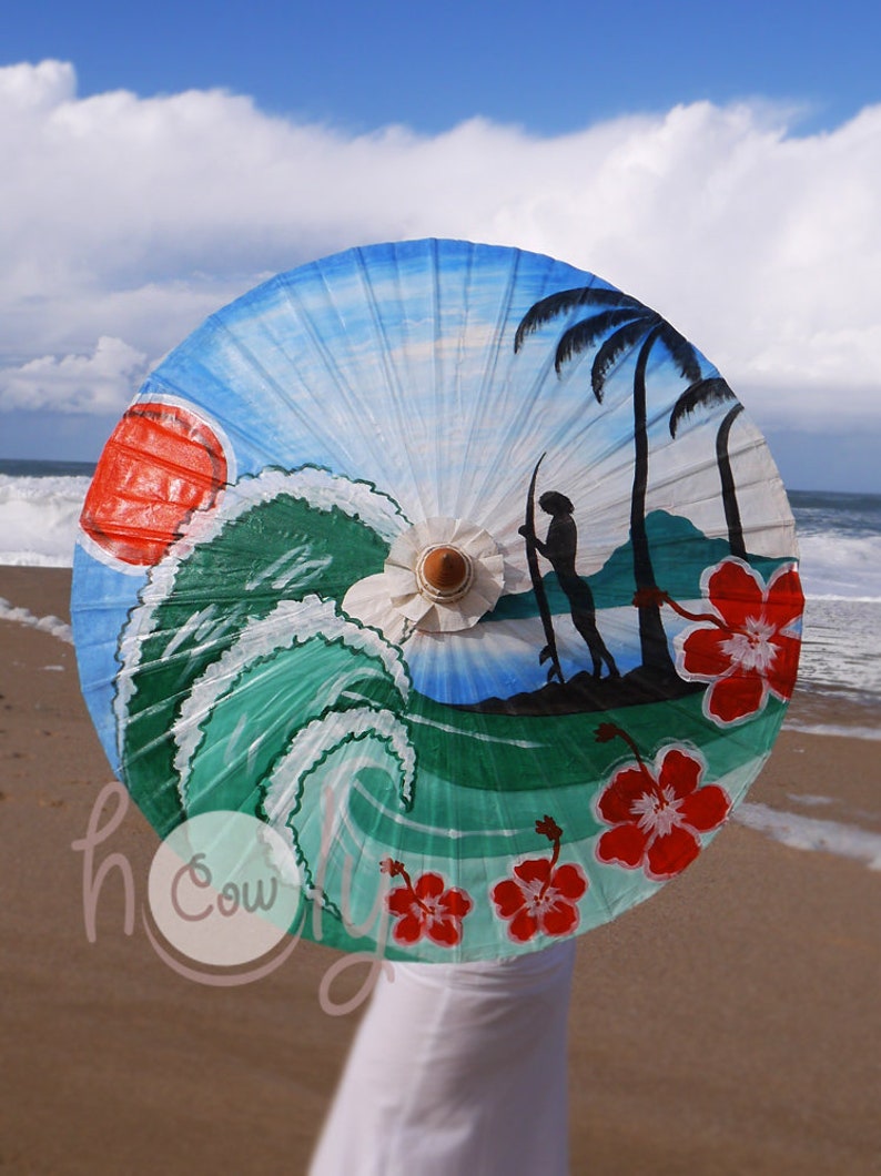 Op de afbeelding: Een handgeschilderde papieren parasol met een strandscene. De parasol heeft een blauwe lucht, een groene golf, palmbomen, een surfer en rode hibiscusbloemen.