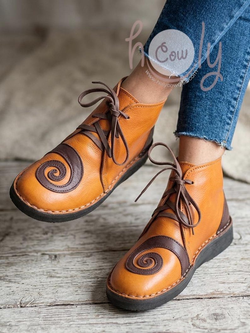 May include: A pair of tan leather ankle boots with brown spiral accents and matching laces. The boots have a dark brown sole and heel, and the text "holy cow" is visible in the background.