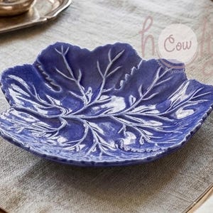 Könnte beinhalten: Eine blaue Keramikschale in Blattform mit weißen Aderdetails. Die Schale steht auf einer neutralfarbenen Leinentischdecke. Das Blattdesign ist geprägt und verleiht dem dekorativen Servierstück Textur.