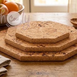 Puede incluir: Tres salvamanteles hexagonales de corcho de diferentes tamaños apilados sobre una mesa de madera. Los salvamanteles son de color marrón claro natural con motas más oscuras. Un cuenco de fruta y cucharas de madera están en el fondo.