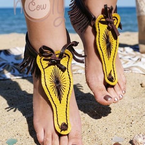 May include: Yellow and brown leather barefoot sandals with a sunburst design. The sandals have a fringe detail and are worn on a sandy beach. The sandals are a unique accessory.