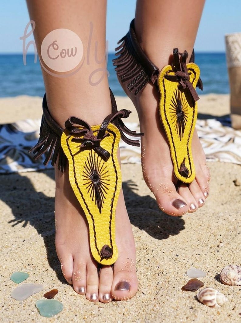 May include: Yellow and brown leather barefoot sandals with a sunburst design. The sandals have a fringe detail and are worn on a sandy beach. The sandals are a unique accessory.