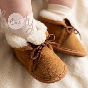 May include: Close-up of two baby booties. The booties are brown suede with a white, fluffy lining and brown laces tied in a bow. The booties are on a soft, neutral-colored surface.