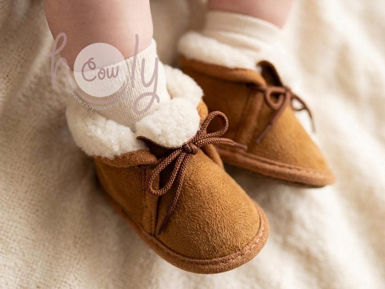 May include: Close-up of two baby booties. The booties are brown suede with a white, fluffy lining and brown laces tied in a bow. The booties are on a soft, neutral-colored surface.