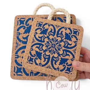 May include: Two square cork coasters featuring a dark blue intricate floral pattern and a natural beige rope handle attached to the top edge. The coasters have rounded corners and a textured surface. The text holy cow chic is visible.