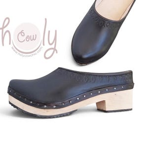 May include: A black leather clog with a light-coloured wooden sole and a low block heel. The upper features a decorative embossed pattern along the collar and is secured to the wooden base with small metal studs. The text holy cow is visible.