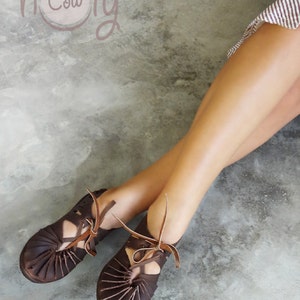 Pu&ograve; includere: Un paio di sandali in pelle marrone con lacci. I sandali hanno un design a taglio e sono indossati da una persona con le gambe nude. Lo sfondo &egrave; una superficie di cemento grigio chiaro. Il testo "holy cow" &egrave; visibile nell'angolo in alto a sinistra dell'immagine.