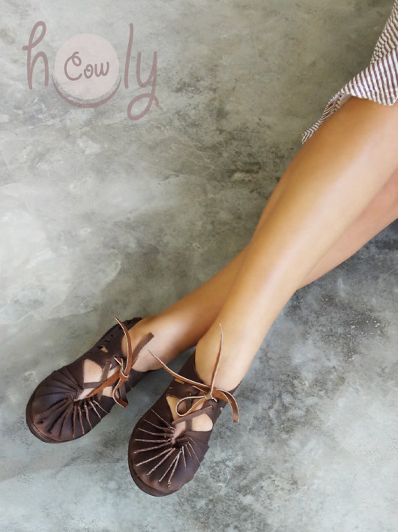 Pu&ograve; includere: Un paio di sandali in pelle marrone con lacci. I sandali hanno un design a taglio e sono indossati da una persona con le gambe nude. Lo sfondo &egrave; una superficie di cemento grigio chiaro. Il testo "holy cow" &egrave; visibile nell'angolo in alto a sinistra dell'immagine.