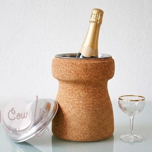 Può includere: Un refrigeratore per vino in sughero con interno in acciaio inossidabile contiene una bottiglia di champagne. Un tappo a forma di cuore e un bicchiere di cristallo con bordo dorato sono vicini. Gli oggetti sono su una superficie di vetro.