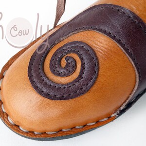 K&ouml;nnte beinhalten: Nahaufnahme eines hellbraunen Lederschuhs mit einem dunkelbraunen Spiraldesign und Akzenten. Der Schuh hat eine gen&auml;hte Sohle und einen Lederriemen. Das Design erinnert an eine Sandale oder einen Pantoffel.